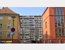 Blick in eine schmale Wohnstraße, eingerahmt von einem gelben Altbau links und einem orangefarbenen Gebäude mit hohen Fensterflächen rechts. Im Hintergrund steht ein mehrstöckiger Plattenbau mit vielen Balkonen; davor sind Straßenlampe, Verkehrsschild, kahle Bäume und parkende Autos zu sehen.