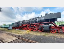 Ausgestellte historische Dampflokomotive in Schwarz und Rot, umgeben von alten grünen Personenwaggons, auf einem Gleisgebiet mit leicht bewölktem Himmel im Hintergrund. Im Vordergrund sind Schienen und eine kleine Sitzbank zu sehen.