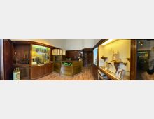 Innenansicht eines kleinen Museums- oder Ladenraums mit dunkler Holzvertäfelung, Vitrinen und Glasregalen mit Exponaten sowie einem zentralen hölzernen Verkaufstresen. Rechts befindet sich eine beleuchtete Wandvitrine mit Ausstellungsobjekten, davor ein Holzboden und im Hintergrund ein Durchgang zu weiteren Räumen.