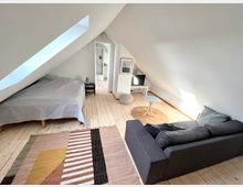 Dachgeschosszimmer mit schrägen weißen Wänden, hellen Holzdielen und einem Dachfenster. Das Zimmer ist mit einem grauen Bett, einem Sofa, einem kleinen runden Tisch, einem Teppich mit geometrischen Mustern und einem Fernseher ausgestattet; im Hintergrund führt eine Tür in ein Badezimmer.