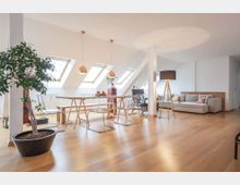 Helles Wohnzimmer im Dachgeschoss mit schrägen Decken und mehreren Dachfenstern, ausgestattet mit einem Esstisch, transparenten Stühlen, einer großen Topfpflanze und einer hellen Couchgarnitur auf einem Holzfußboden. Kupferfarbene Pendelleuchten und eine Stehlampe sorgen für die Beleuchtung.