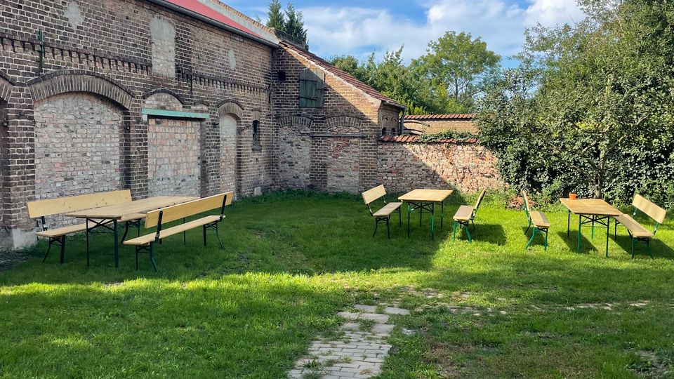 Innenhof mit Rasenfläche und schmalem gepflastertem Weg, umgeben von alten Backsteinmauern mit zugemauerten Bögen und einem Gebäude mit rotem Dach. Auf der Wiese stehen mehrere einfache Holzbänke und Tische, daneben Bäume und Sträucher am Rand.