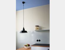 Minimalistisch eingerichtete Küche mit cremefarbenen Wänden und einer blauen Decke. Eine schwarze Hängelampe, eine Schale mit Äpfeln auf der Holzarbeitsplatte und ein kleines Regal mit einer goldenen Waage prägen den Raum.