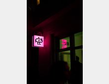 Fassade eines Gebäudes bei Nacht mit einem leuchtenden, pinken Schild, das ein stilisiertes Symbol zeigt. Durch die Fenster ist grünliches Licht sichtbar, und im Vordergrund befinden sich unscharfe Silhouetten von Personen.