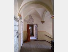 Ein renovierungsbedürftiger Gebäudeflur mit spitzbogigen Kreuzgewölben, einer sichtbaren Wandbeschädigung, verblassten gelblichen Tapeten und einem roten Tuch im Türrahmen. Am rechten Rand führt eine schmale Treppe mit Holzhandlauf nach unten.