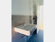 Ein modernes Badezimmer mit einem rechteckigen Waschbecken, grauen Wandfliesen und blau gemusterten Bodenfliesen. Rechts neben dem Waschbecken befindet sich ein weißer Handtuchheizkörper, und eine geöffnete Tür führt zu einem Raum mit hellem Holzboden.