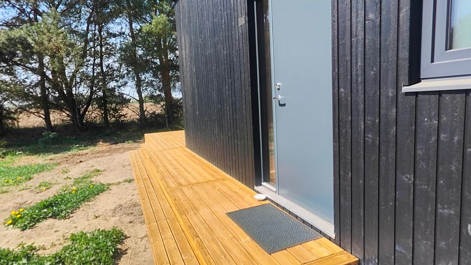 Schwarzes Holzgebäude mit vertikaler Verschalung, grauer Eingangstür und Fenster an der Seite, davor eine kleine Holzterrasse mit Stufe und Fußmatte. Das Gebäude steht in einer sandigen, spärlich bewachsenen Umgebung mit Bäumen im Hintergrund unter blauem Himmel.