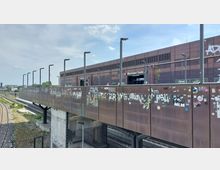 Bahnhof Warschauer Straße in Berlin, mit einer erhöhten Fußgängerbrücke aus Metall, die über Bahngleise führt. Die Brücke und das Bahnhofsgebäude sind mit Graffiti und Aufklebern bedeckt, im Hintergrund ist eine Bahnsteigszene sichtbar.