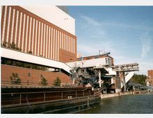 Industrieanlage mit Backsteinfassaden und großen rechteckigen Fenstern, angrenzend an einen Wasserkanal. Im Vordergrund ist ein Frachtschiff zu sehen, daneben Förderbänder und metallene Strukturen, die die Anlage verbinden.