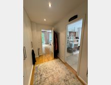 Flur einer Wohnung mit hellem Holzboden und eingebauten weißen Schränken auf der linken Seite. Der Flur führt zu einem Wohnzimmer mit Fenstern und Vorhängen sowie einem Badezimmer mit gefliesten Wänden und Glasdusche auf der rechten Seite.