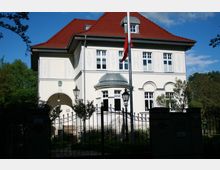 Zweistöckige Villa mit heller Fassade und rotem Satteldach, großen Fenstern und einem kleinen Erker mit Kuppeldach. Im Vordergrund ein schmiedeeisernes Tor, flankiert von Laternen und umgeben von Bäumen und Sträuchern.