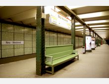 U-Bahn-Station „Neu-Westend“ in Berlin mit grünen Metallstützen, einer grünen Sitzbank im Vordergrund und gefliesten Wänden im Hintergrund. Die Station verfügt über ein schlichtes Design mit Leuchtstoffbeleuchtung und Werbesäulen entlang des Bahnsteigs.