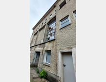 Ein mehrstöckiges Gebäude mit grober Putzfassade, kleinen rechteckigen Fenstern und einem großen Fenster mit roten Metallrahmen in der Mitte. Im Vordergrund ein einfacher Zaun, eine Metalltür und etwas Vegetation am unteren Rand der Fassade.