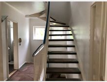 Treppenhaus in einem Wohngebäude mit offener Holztreppe und Metallgeländer, einem Fenster mit Blick ins Grüne sowie Zugang zu einem Badezimmer mit weißen Fliesen. Die Wände sind weiß gestrichen, und der Boden ist teilweise mit einer Schutzmatte bedeckt.