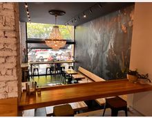 Innenraum eines modernen Cafés mit Holzmöbeln, einer großen Fensterfront und einem großen Kronleuchter. Die Wände sind mit gemusterten Tapeten und weißer Ziegeloptik gestaltet, und durch das Fenster sind geparkte Autos sowie ein Gehweg mit Passanten sichtbar.