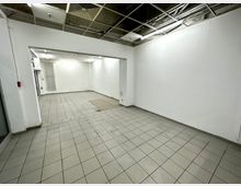 Leerer Innenraum mit gefliesten Böden und weißen Wänden, Decke teilweise mit sichtbaren Kabeln und Lüftungselementen. Im Hintergrund ein Heizkörper und eine beleuchtete Nische.