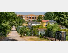 Schotterweg durch eine grüne, dicht bewachsene Wohngegend mit Gärten und vereinzelten Einfamilienhäusern. Im Hintergrund ein größeres, rötliches Mehrfamilienhaus und Bäume, die den Horizont umrahmen.