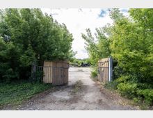 Offenes Metalltor mit Holzverkleidung, das zu einem verlassenen, teilweise bewachsenen Gelände mit Betonboden führt; dichte grüne Büsche und Bäume säumen den Eingang. Im Hintergrund sind gestapelte Reifen zu erkennen.