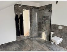 Modern bathroom with large gray stone-look tiles, a walk-in glass shower with rainfall showerhead and recessed shelf, and a wall-mounted toilet. A tall mirror and towel hooks are on the left wall, with white upper walls and small ventilation grilles near the ceiling.