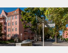 Straßenecke in einer Wohngegend mit rosa Putzgebäuden im historisierenden Stil, Bäumen entlang der Straße und Straßenschildern mit den Namen "Eisenacher Straße" und "Lerchenweg". Im Vordergrund sind geparkte Autos, ein Gehweg und ein Fußgänger zu sehen.