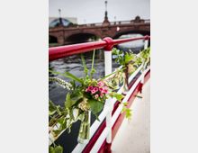 Ein geschmücktes Geländer auf einem Boot mit Blumen in kleinen Glasvasen, im Hintergrund eine historische Backsteinbrücke über einen Fluss. Die Szene spielt sich unter freiem Himmel ab.
