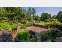 Steingarten mit gewundenen Wegen aus Naturstein, umgeben von niedrigem, mit Pflanzen bewachsenem Mauerwerk, blühenden Beeten und großen Bäumen im Hintergrund. Die Szenerie zeigt eine gepflegte Grünanlage unter klarem Himmel.