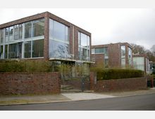 Moderne Wohngebäude mit ziegelverkleideter Fassade und großen Glasfronten, umgeben von einer niedrigen Backsteinmauer und Hecken. Im Vordergrund ein Eingangstor mit gepflastertem Weg, angrenzend an eine ruhige Straße.