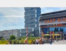 Moderne Einkaufs- und Bürogebäude mit einem geschwungenen Glasfrontbereich, beschriftet mit „East Side“, daneben ein hoher, gestaffelter Büroturm. Im Vordergrund sind Fahrräder geparkt, Passanten und Sonnenschirme eines Außenbereichs zu sehen, umgeben von Bäumen.
