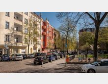 Straßenszene in einer Wohngegend mit mehrstöckigen Altbauwohnungen, darunter ein auffälliges rotes Gebäude. Im Vordergrund sind parkende Autos, ein Gehweg, sowie ein kleiner Park mit Bäumen und einem Spielplatz zu sehen.