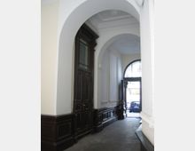 Eingangsbereich eines historischen Gebäudes mit einem hohen, gewölbten Durchgang. Wände sind cremefarben, mit dunklem Holzvertäfelung und einer großen Tür aus dunklem Holz; im Hintergrund ist eine offene Glastür, die den Blick nach draußen auf parkende Autos freigibt.