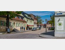 Straßenszene in einer historischen Altstadt mit gepflastertem Gehweg, farbig bemalten Gebäuden mit Satteldächern, grünen Fensterläden und kleinen Geschäften. Im Vordergrund gehen Menschen entlang der Straße, parkende Autos und Laternen säumen den Bereich, während im Hintergrund Bäume und ein klarer Himmel zu sehen sind.