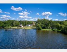 Seeufer mit klarem, blauem Wasser, einer Anlegestelle und einer bewaldeten Uferlandschaft im Hintergrund. Mehrere Gebäude, darunter ein gelbes Haus mit Turm, stehen umgeben von grünen Bäumen unter einem Himmel mit einzelnen weißen Wolken.
