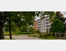 Mehrstöckiges Wohngebäude mit beige und orangefarbenen Fassaden, umlaufenden Balkonen und umgeben von Bäumen und Sträuchern. Im Vordergrund ein unbefestigter Weg, Wiesenflächen und ein Zaun.