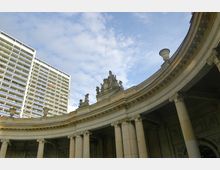 Halbkreisförmiger Gebäudebogen mit säulengetragener Fassade und dekorativen Skulpturen auf dem Dach, im Hintergrund ein modernes Hochhaus vor einem blauen Himmel mit Wolken.