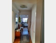 Ein schmaler Flur in einem Wohnhaus mit Holzboden, einem kleinen Teppichläufer, einer Kommode an der linken Wand und einer Treppe auf der rechten Seite. Im Hintergrund ist ein Raum mit einem Fenster und einem Leuchtenschirm aus farbigem Glas sichtbar.