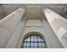 Blick von unten auf eine Fassade mit massiven Säulen aus Stein, einem großen Rundbogenfenster und einer Inschrift mit der römischen Zahl "MDCCLII" in Gold. Die Architektur weist auf ein historisches Gebäude mit klassizistischen Elementen hin.