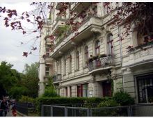 Mehrstöckiges Wohngebäude im Stil der Gründerzeit mit verzierten Fassaden, roten Fensterrahmen und dekorativen Balkonen, die mit Blumen geschmückt sind. Im Vordergrund befinden sich ein Gehweg, ein Zaun, sowie Bäume und Sträucher.