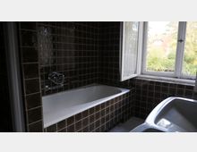 Badezimmer mit dunklen Fliesen, einer weißen Badewanne und einer offenen Fensterfront mit Blick ins Grüne. Links der Badewanne sind verchromte Armaturen angebracht, und der Boden ist mit kleinen Mosaikfliesen bedeckt.