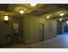 Eingangshalle mit grobkörnigen Wänden, einer beleuchteten Kassettendecke und mehreren Lampen. An der Wand befinden sich ein Münztelefon mit Graffiti sowie eine graue Tür mit Verbotsschildern, daneben ist ein dunkler Metallzaun sichtbar.