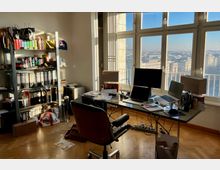 Ein Büro mit großen Fenstern, die einen Ausblick auf eine Stadtlandschaft bei Tageslicht bieten. Der Raum ist mit einem Schreibtisch mit Arbeitsmaterialien, mehreren Laptops und Monitoren sowie einem Bücherregal mit Ordnern und Büchern ausgestattet.