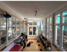 Ein heller Flur mit Holzboden und weißen Wänden, gesäumt von großen Fenstern auf beiden Seiten. Entlang der Wände stehen Holzregale voller Schuhe, und am Ende des Flurs befindet sich eine kleine Fensterfront mit Blick auf einen Außenbereich.