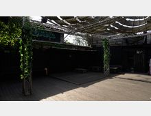 Eine überdachte Außenfläche mit Holzboden, umgeben von schwarzen Containern und dunklen Wänden. Metallgerüste mit Kletterpflanzen und eine Wellblechüberdachung aus Stoff sorgen für Schatten, während vereinzelte Palettenmöbel den Raum einrahmen.