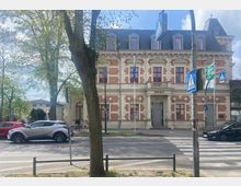 Historisches Rathausgebäude aus rotem Backstein und hellem Stein mit symmetrischen Fenstern und einem Eingang mit Säulen. Im Vordergrund eine Straße mit Zebrastreifen, Verkehrsschildern und parkenden Autos; Bäume rahmen die Szene ein.