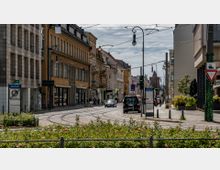 Straßenszene in einer Innenstadt mit historischen Gebäuden, Straßenbahn-Schienen und einer Mischung aus Fußgängern und Fahrzeugen. Im Vordergrund befinden sich Beete mit blühenden Rosen, während im Hintergrund eine Kirche mit Turm sichtbar ist.