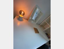 Ein minimalistischer Schreibtisch mit heller Oberfläche, einer leuchtenden Tischleuchte mit Filament-Design, einem Rahmen mit Kunstmotiv und einem geschlossenen Notizbuch. Rechts daneben befinden sich weiße Regalfächer mit Stiften und Büchern.