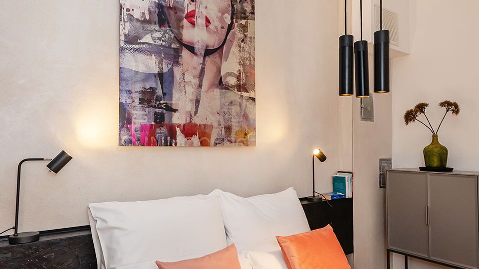 Modernes Schlafzimmer mit Doppelbett in weißer Bettwäsche und zwei korallfarbenen Kissen, flankiert von kleinen Nachttischlampen. Über dem dunklen Kopfteil hängt ein großes, farbiges Kunstbild; von der Decke hängen drei zylindrische Pendelleuchten, daneben steht ein Sideboard mit Vase.