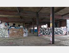 Industriehalle mit sichtbarer Beton- und Stahlkonstruktion, deren Wände mit buntem Graffiti bedeckt sind. Der Boden ist aus Rohbeton, und die Umgebung wirkt verlassen und ungenutzt.