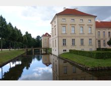 Heller, klassizistischer Schlossbau mit roten Ziegeldächern, umgeben von einem Wassergraben und gepflegtem Rasen. Eine kleine Brücke führt über den Wassergraben, links sind Bäume und ein Weg mit Spaziergängern zu sehen.