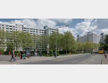 Eine Wohnsiedlung mit mehreren großen, rechteckigen, weißen Mehrfamilienhäusern und zahlreichen Fenstern. Im Vordergrund eine Bushaltestelle mit Personen, umgeben von Bäumen und Gehwegen, sowie eine Straße mit einem geparkten Linienbus.