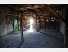 Verlassener Dachboden mit Holzbalkenkonstruktion, Holzboden und einer grünen Tür an einer Ziegelsteinwand. Das Dach ist teilweise gedeckt, und Lichtstrahlen dringen durch Lücken in die dunkle, staubige Umgebung ein.